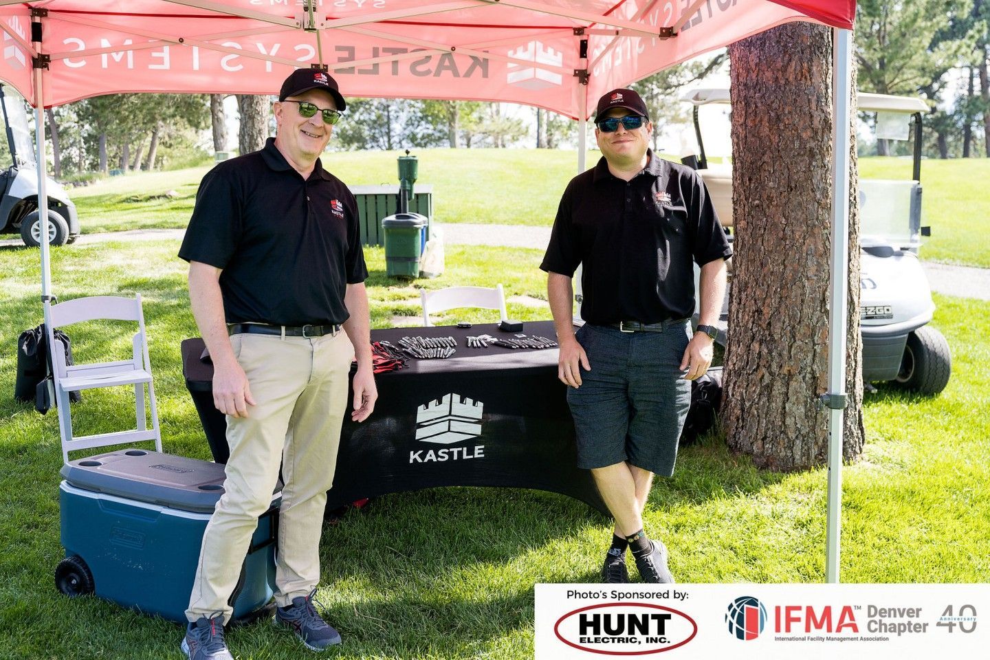 Two men standing under a branded canopy at a golf event. One wears khaki pants, the other shorts.
