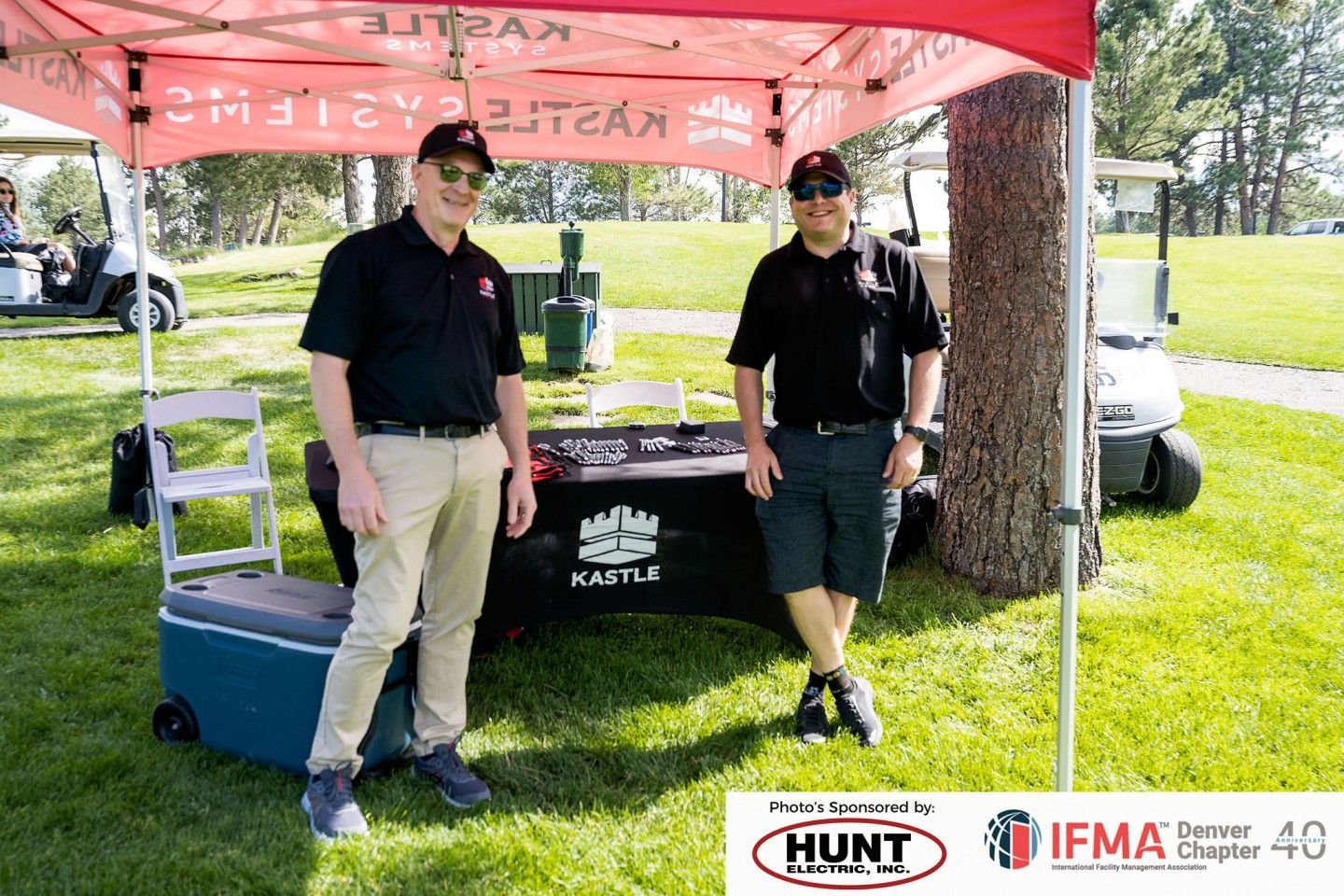 Two men stand under a red tent with a table at an outdoor event. One wears khakis; the other shorts.