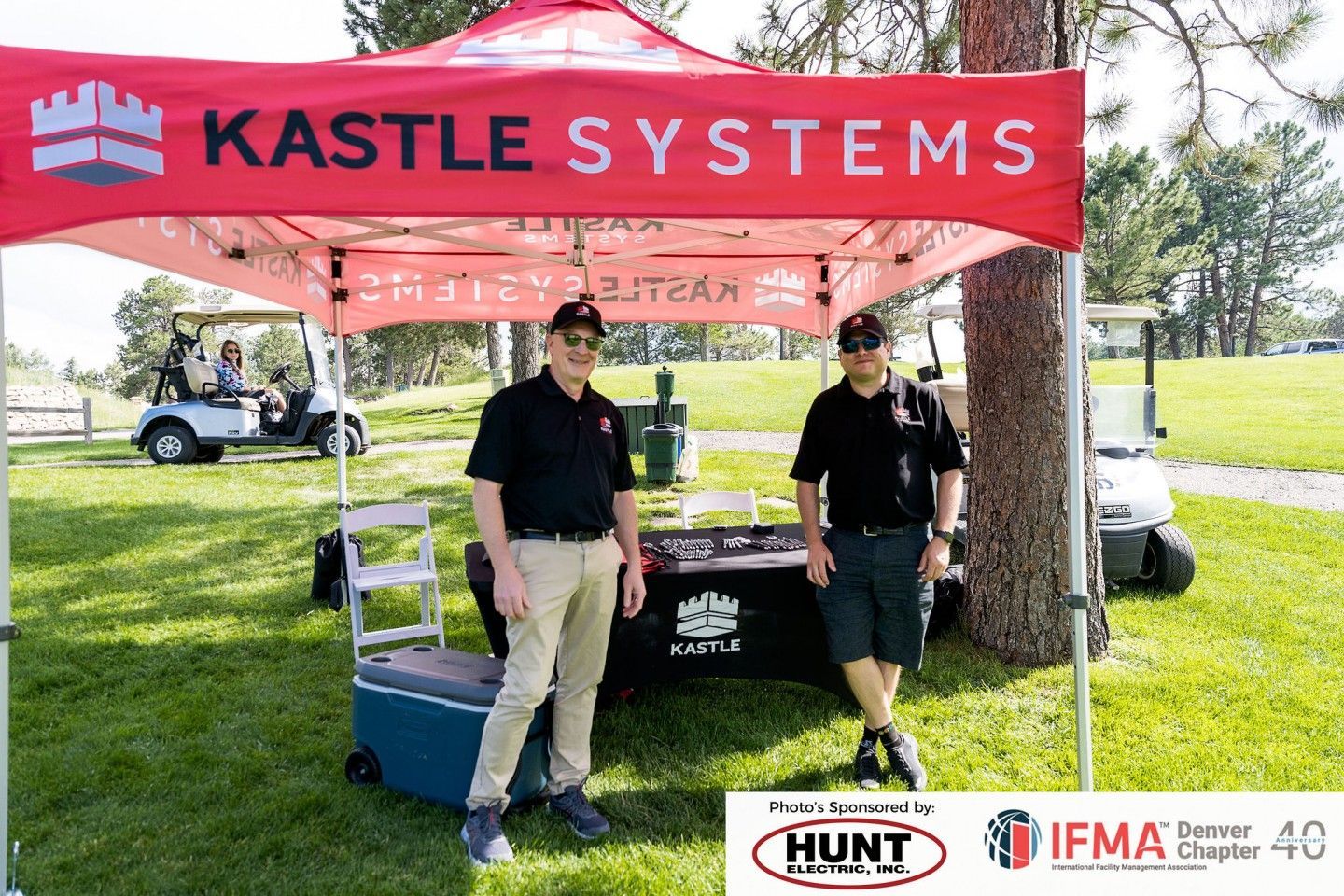 Two men stand at a Kastle Systems booth at an outdoor event, beside a table, tent, and golf carts.