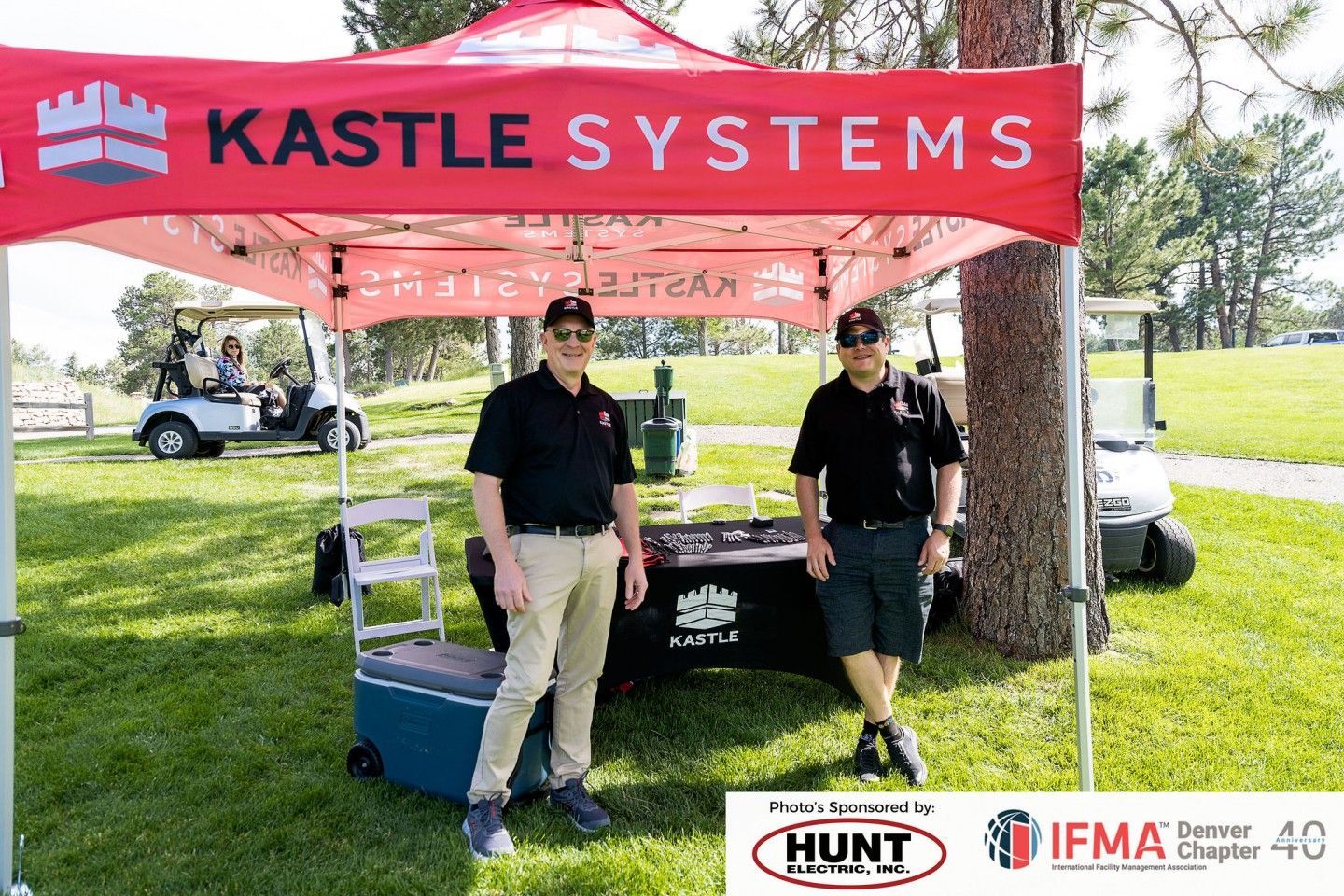 Two men under a Kastle Systems tent at a golf event; one in khaki pants, one in shorts.