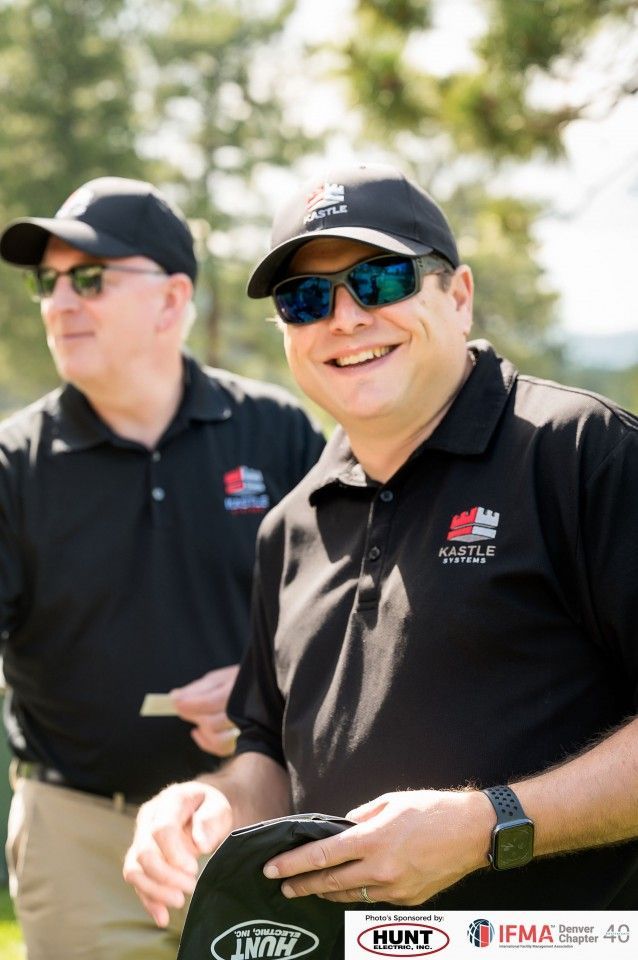 Two men in black shirts and hats on a golf course; one smiles.