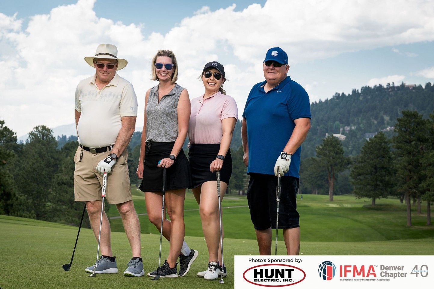 Four people on a golf course holding clubs; sunny day, mountains in the background, event logo visible.