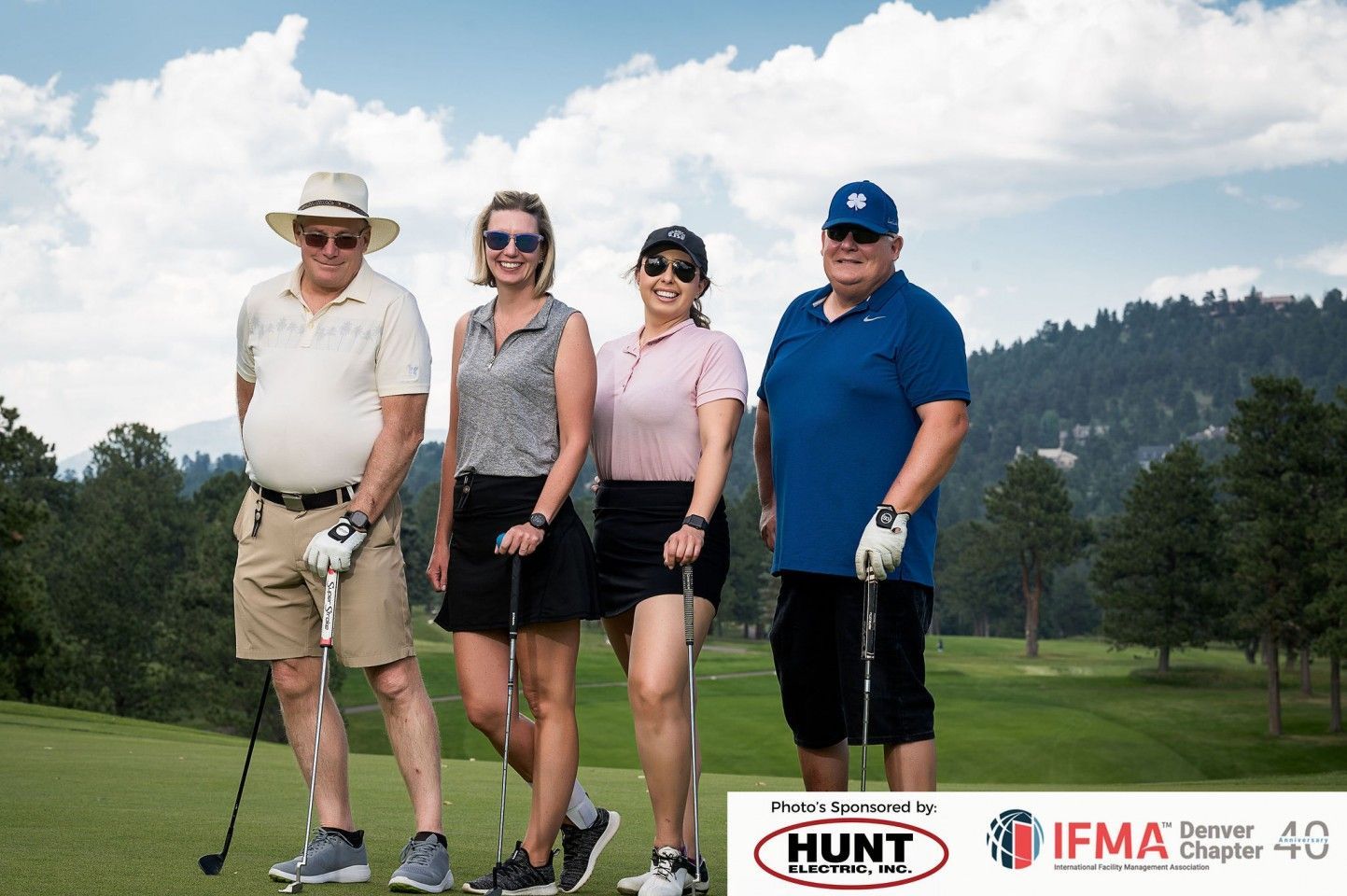 Four people on a golf course. They stand with golf clubs. Mountains are in the background. Sunny day.