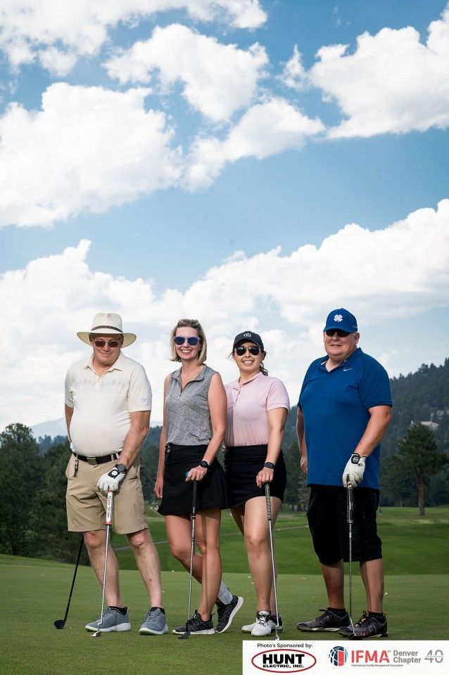 Four people posing on a golf course under a cloudy sky. Each holds a golf club.