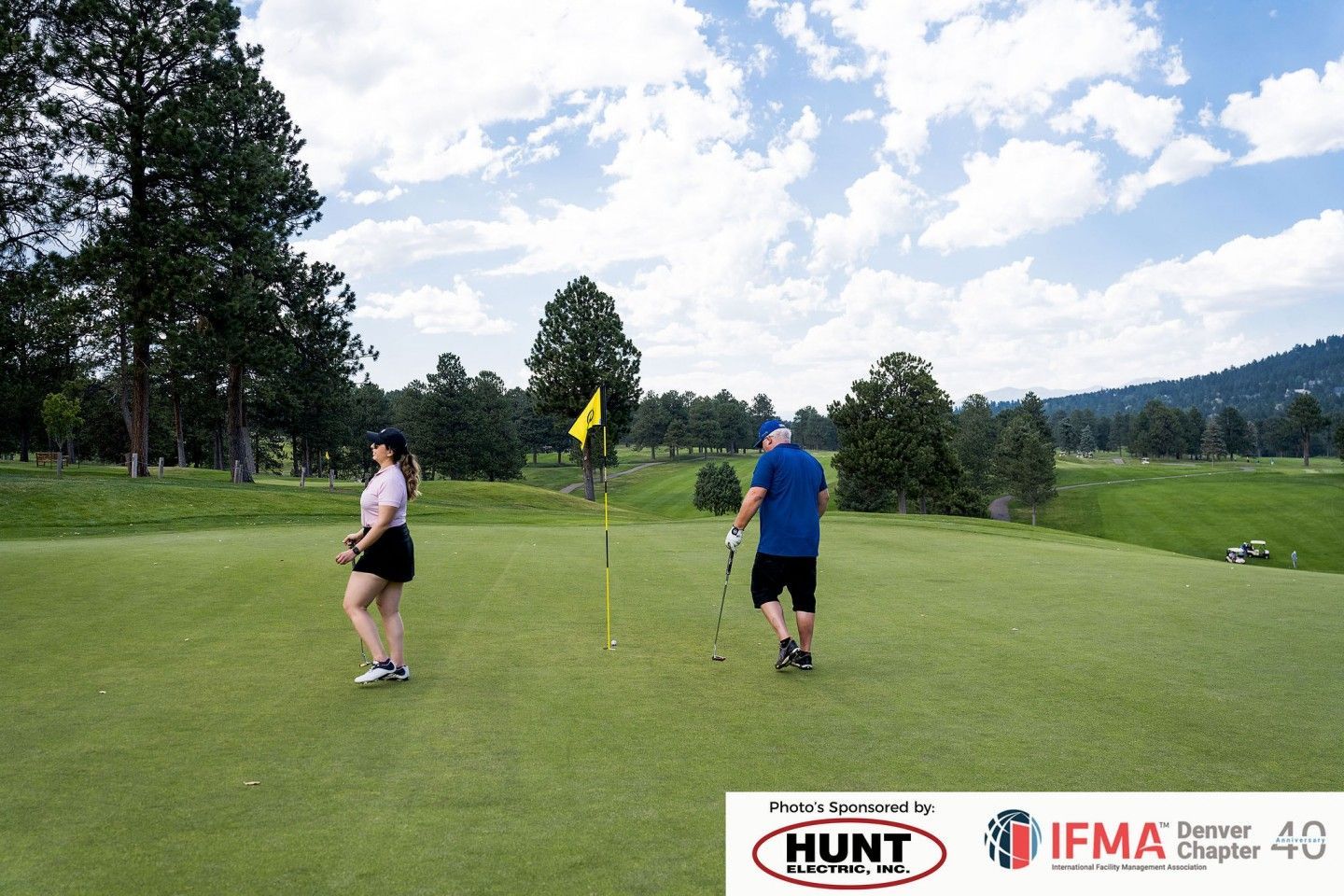 Two people on a golf green with a flag. One in pink shirt and cap, the other in blue shirt, sunny day.