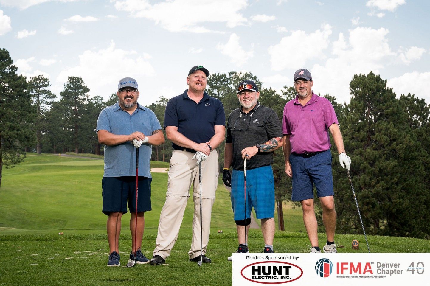 Four men on a golf course holding clubs, smiling. Sunny day, green grass, trees in background.