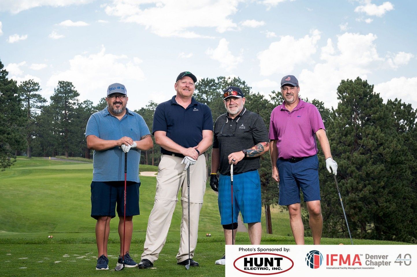 Four men in golf attire pose on a golf course. Each holds a club, smiling, with a logo in the corner.
