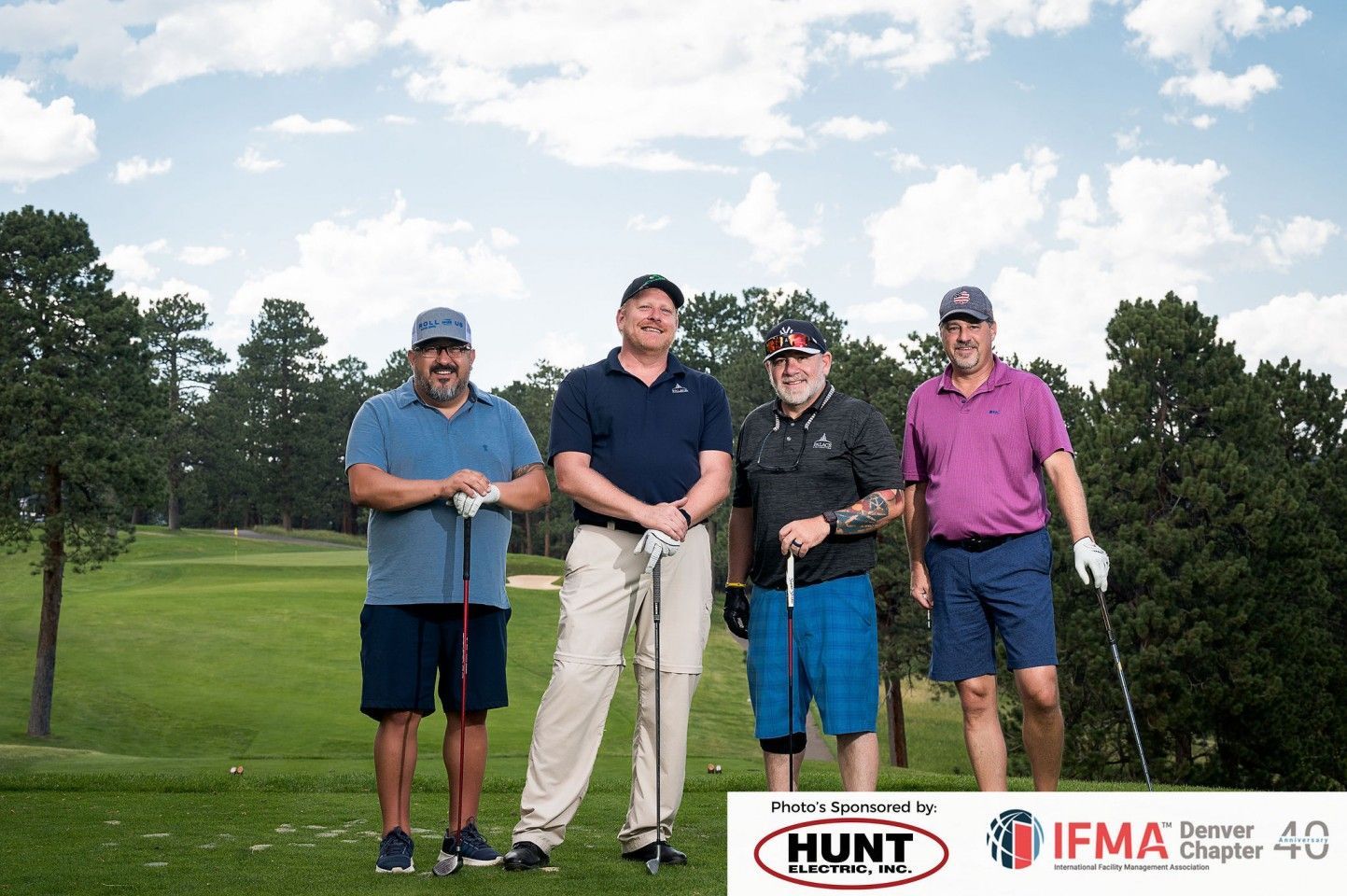 Four golfers on a green golf course, smiling and posing. Sponsors' logos are on the bottom.