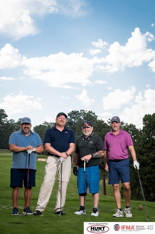 Four men on a golf course pose with clubs. Sunny day, green grass, blue sky.