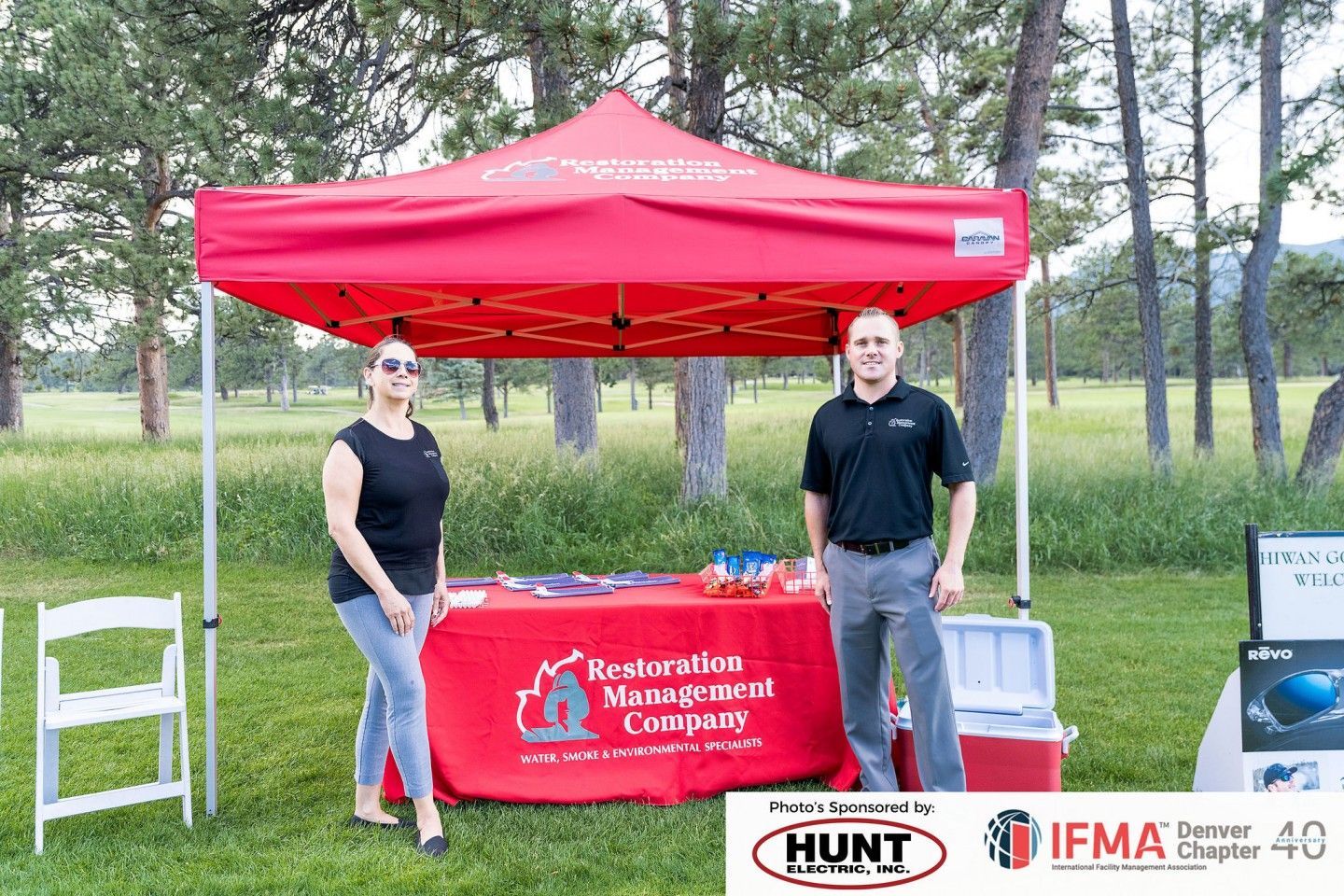Two people at a Restoration Management Company booth under a red canopy. Setting is outdoors on grass with trees.