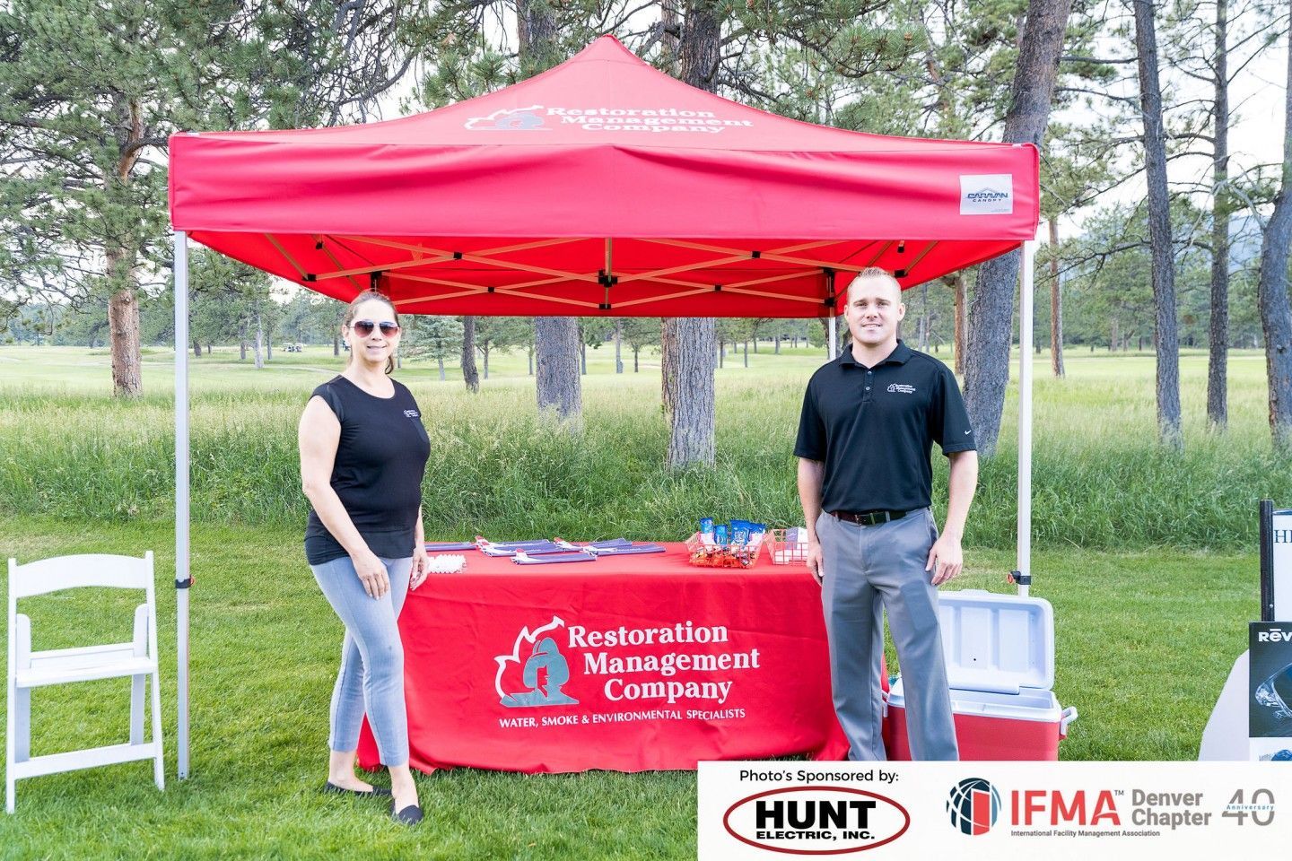 Two people stand by a red tent with a company logo. They are outdoors on grass, with trees in the background.