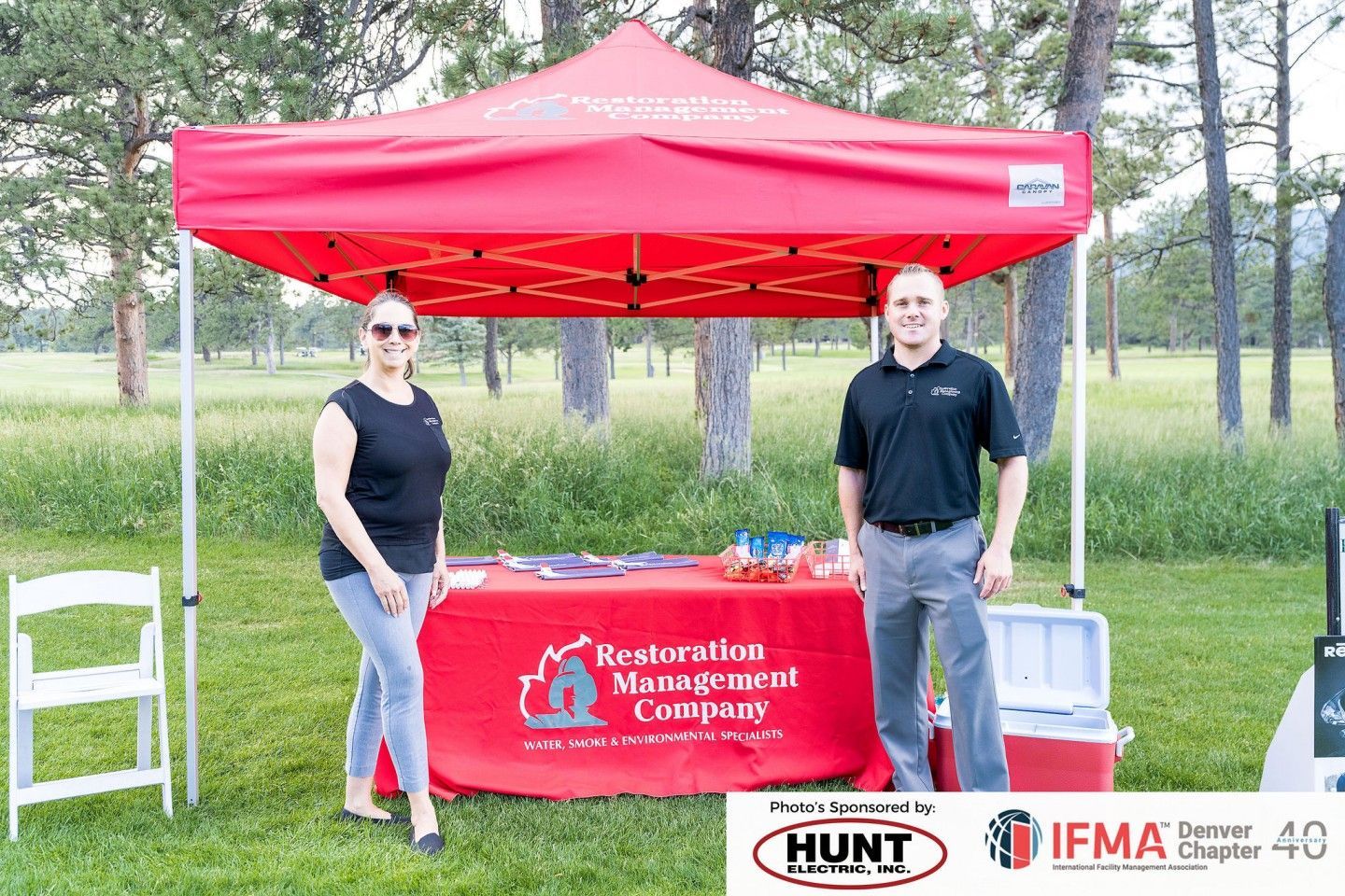 Two people stand behind a red tent with a company logo.