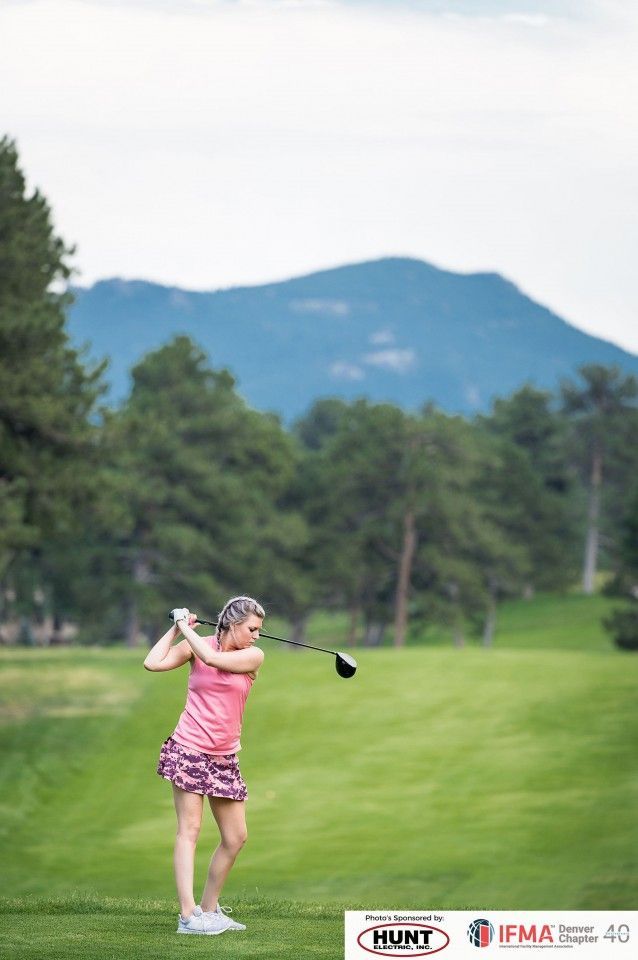 Woman in pink golf attire swings a club on a green course with mountains in the background.