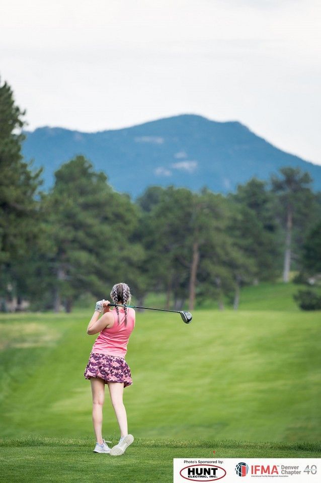Golfer swings a club on a green course with mountains and trees in the background. She wears pink and purple attire.