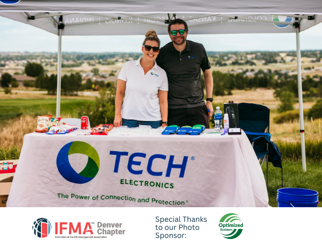 Two people at a TECH Electronics booth at an outdoor event. A table displays snacks.