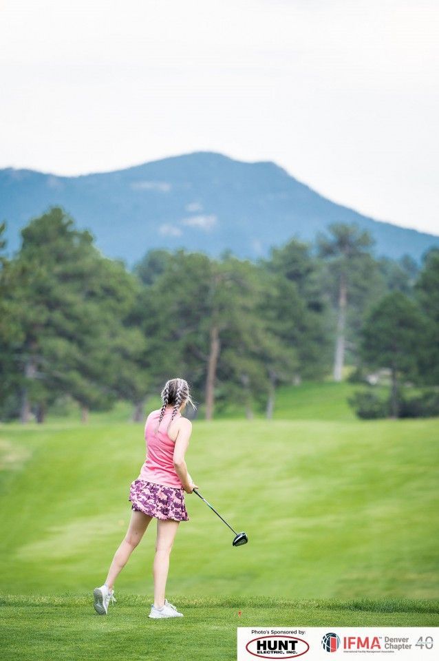 Woman in pink golf attire swinging a club on a green course with mountains in the background.