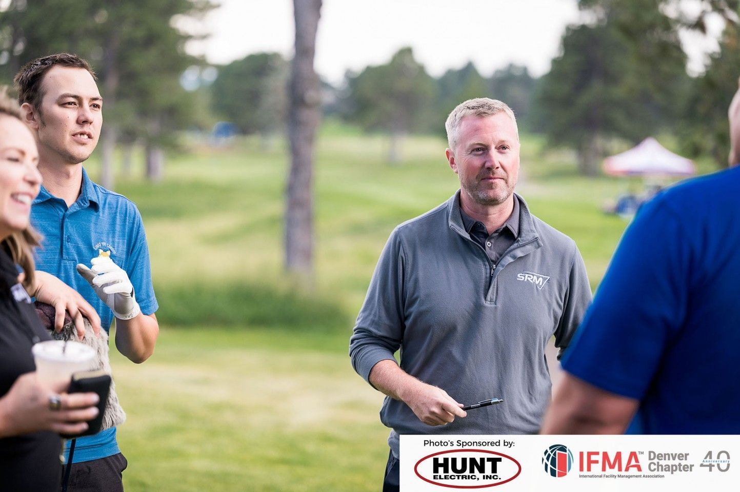 Group of people talking on a golf course. Man in gray shirt holds a pen, others hold drinks, and the sky is overcast.