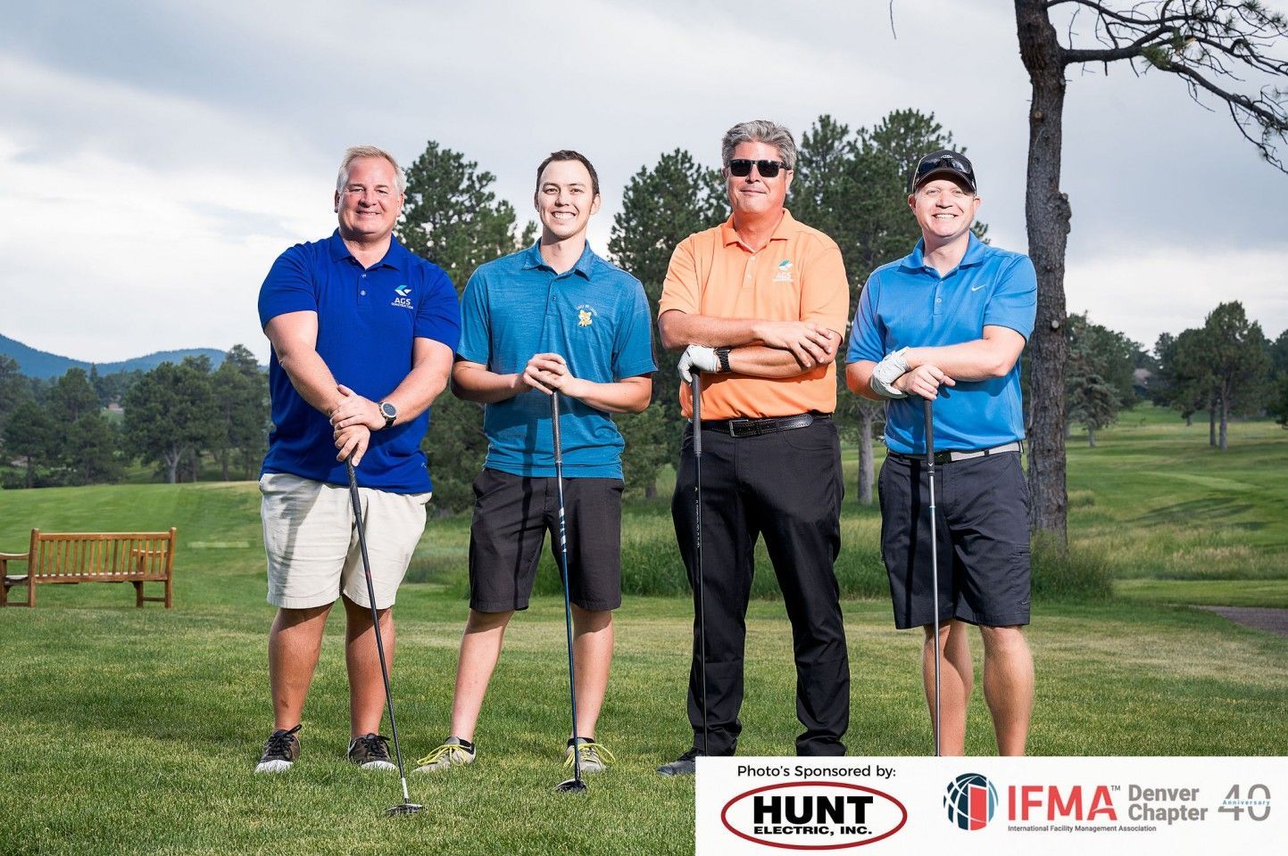 Four men with golf clubs on a course; one in an orange shirt.
