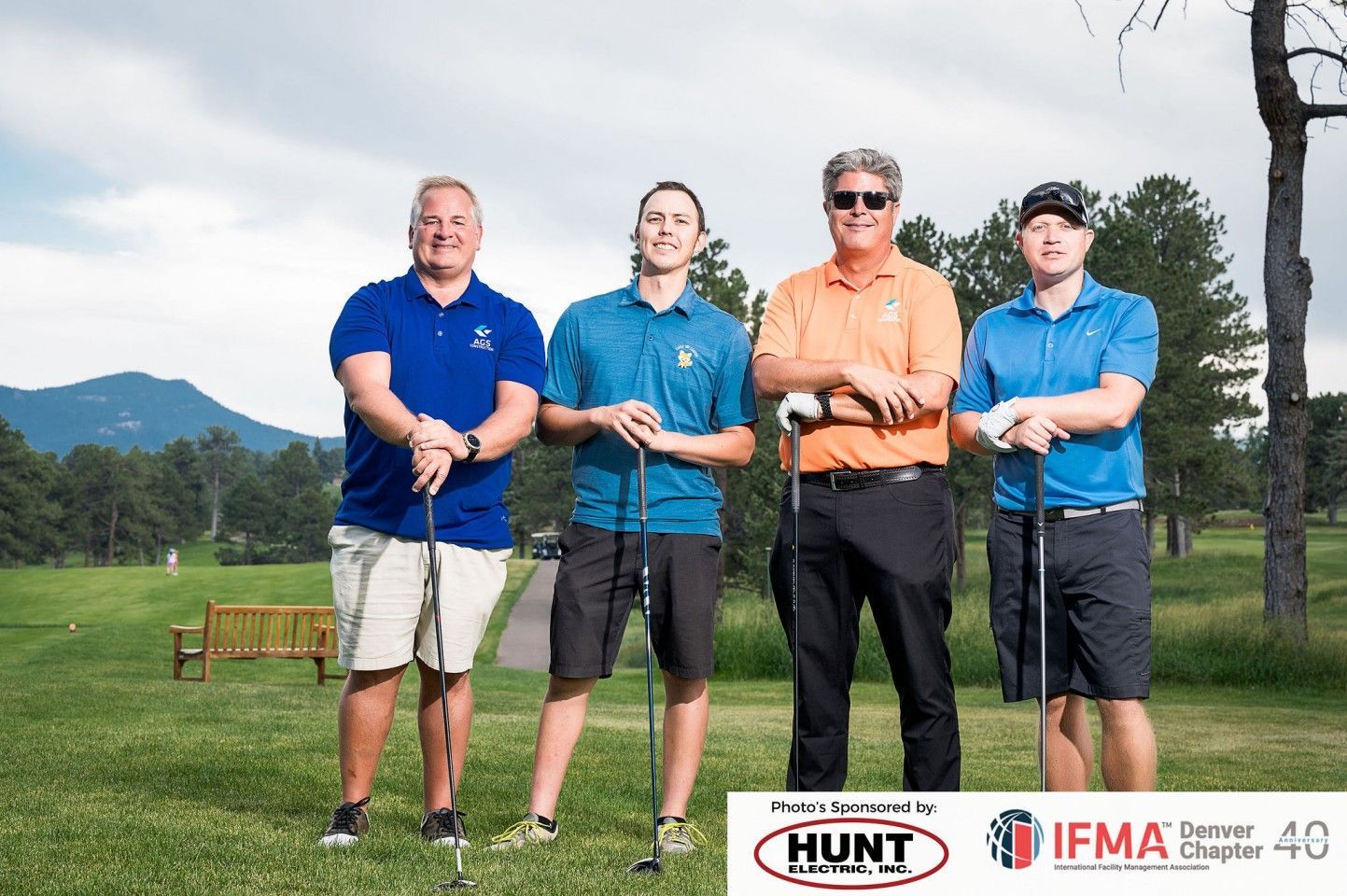 Four people with golf clubs standing on a golf course. They wear casual shirts and shorts.