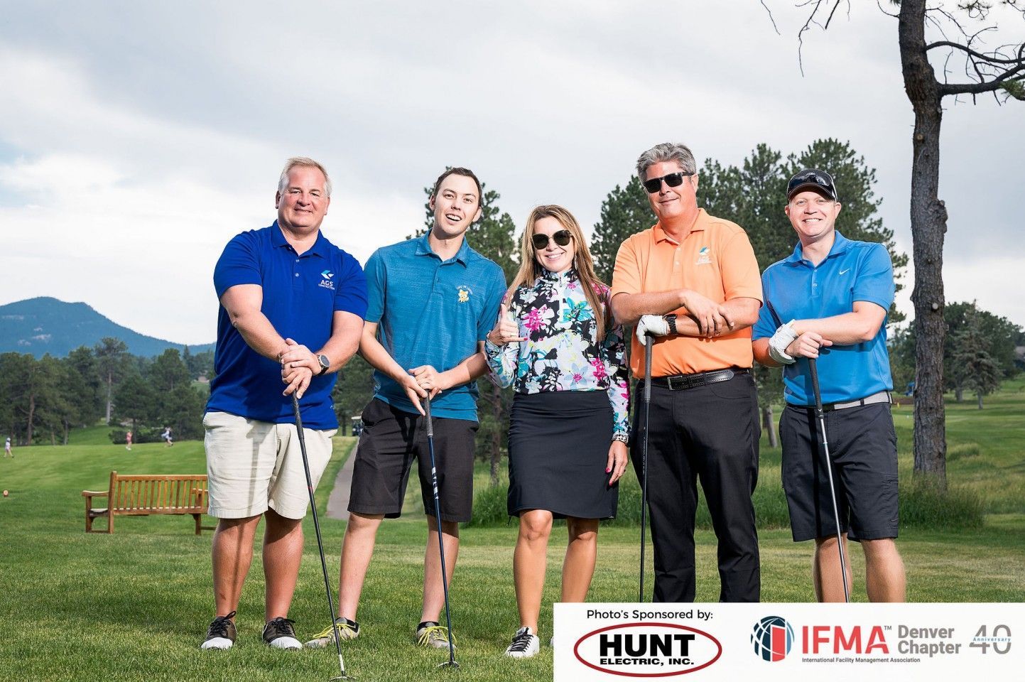 Five people holding golf clubs on a golf course; one woman in the middle, smiling.