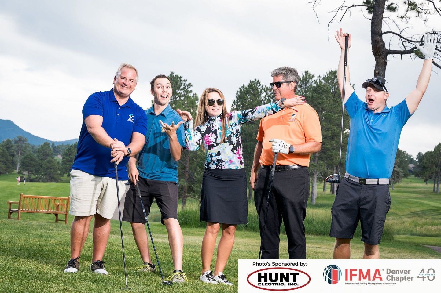 Five people on a golf course celebrating, arms raised, mixed expressions. Mountains, trees, and sky in the background.