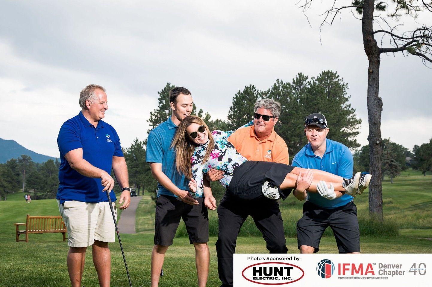 Group of five people on a golf course. Four men holding a woman playfully; one man watches, smiling.