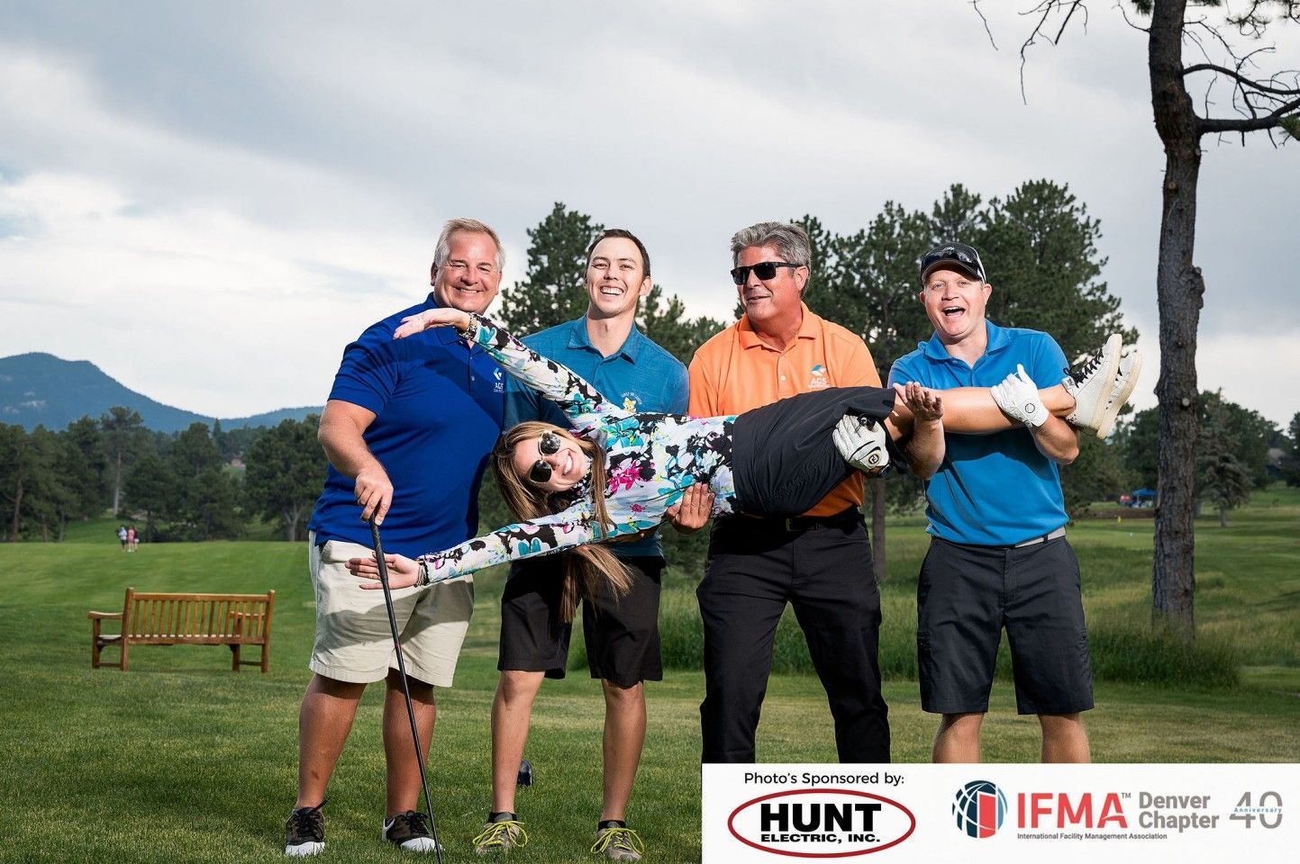 Four people, two holding up a person, laughing on a golf course with mountains in the background.