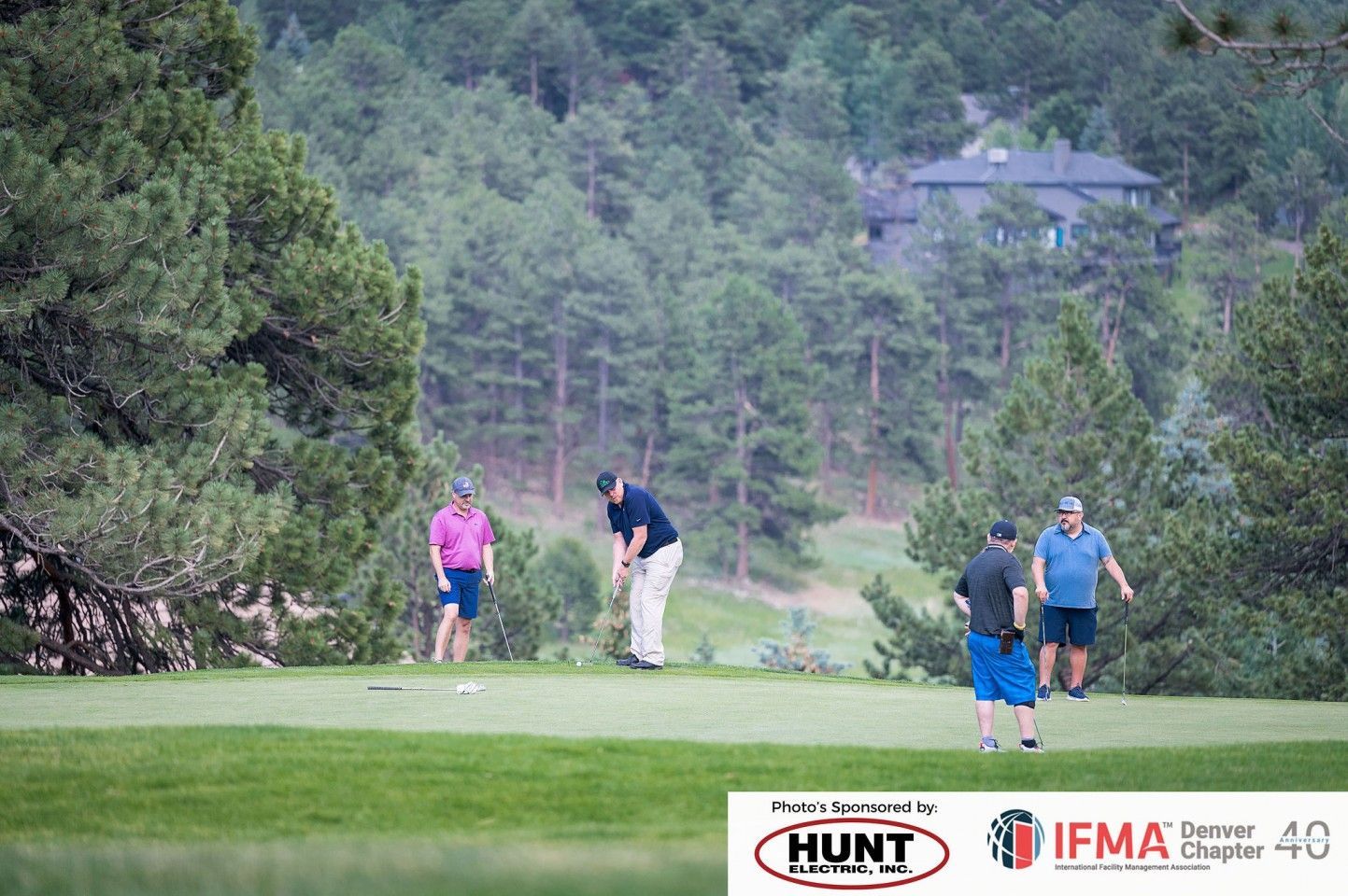 Golfers putting on a green, surrounded by trees and a house in the background.