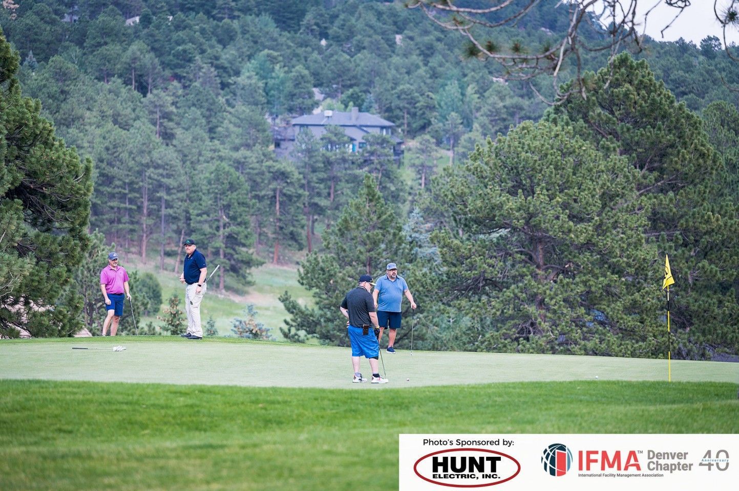 Golfers on a green, trees and a building in the background. Two put, two look on.