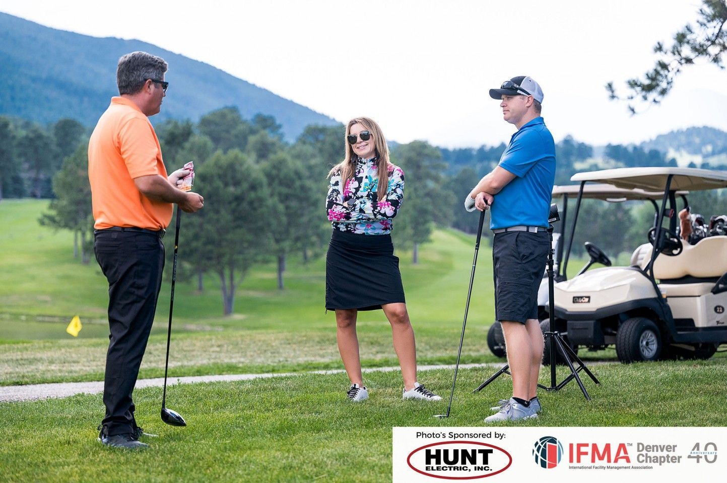 Three people on a golf course. A man in orange shirt and another in blue shirt converse with a woman in floral top.