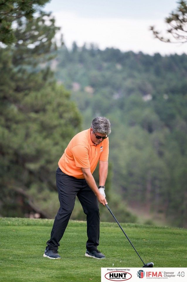 Man in orange shirt swings golf club on a green course; trees in background.
