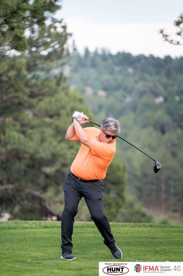 Man in orange shirt swings golf club on a green course, with trees in the background.