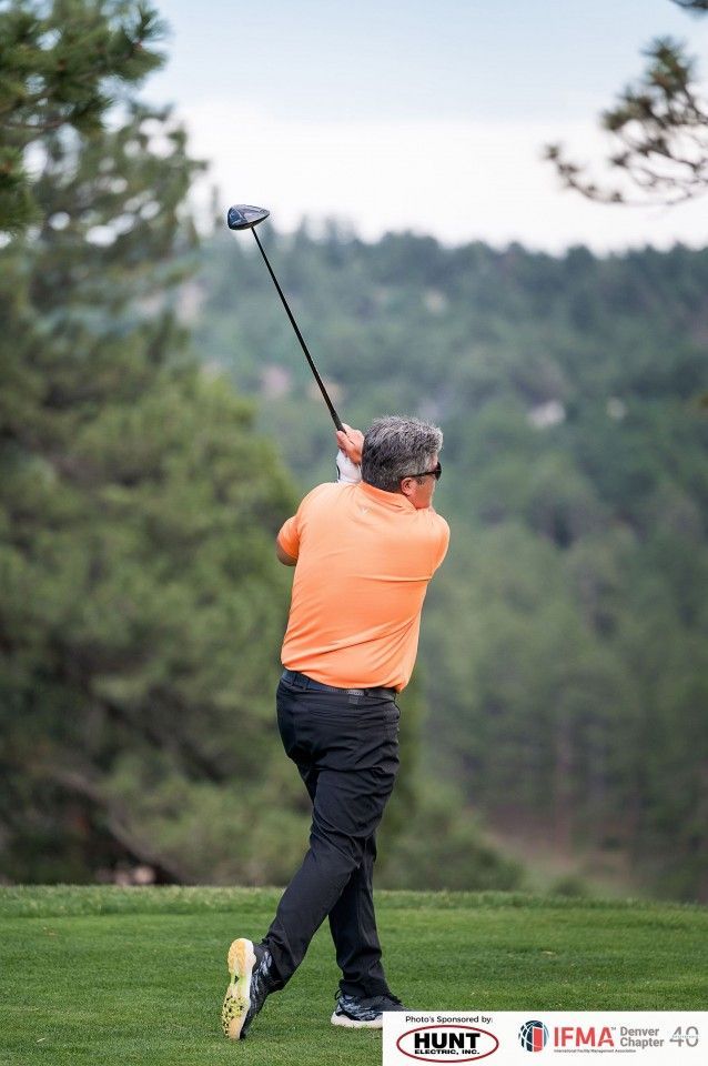 Man in orange shirt swinging a golf club on a green course, with trees in the background.