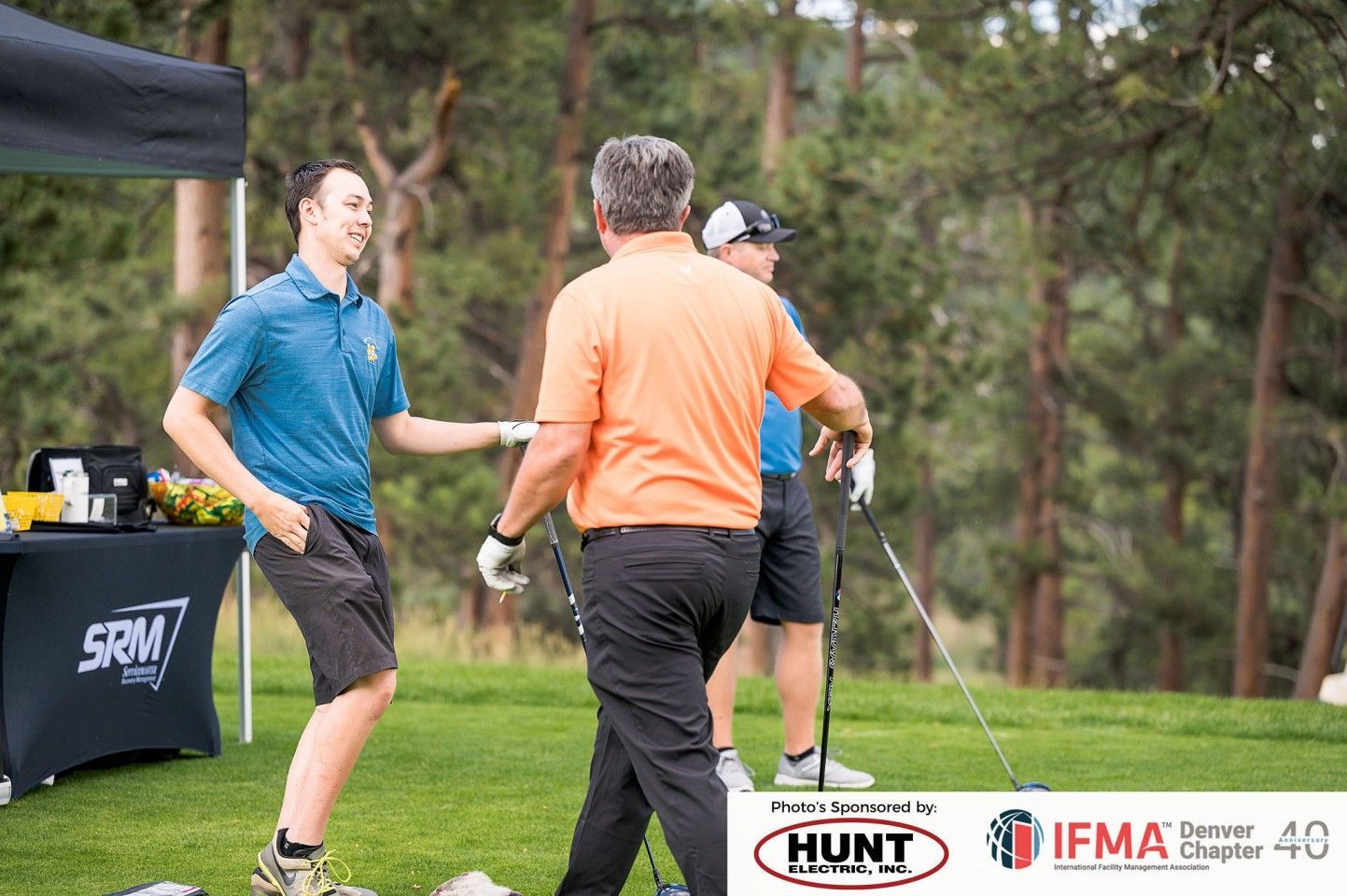 Men on a golf course: two shaking hands, one wearing orange, another blue.
