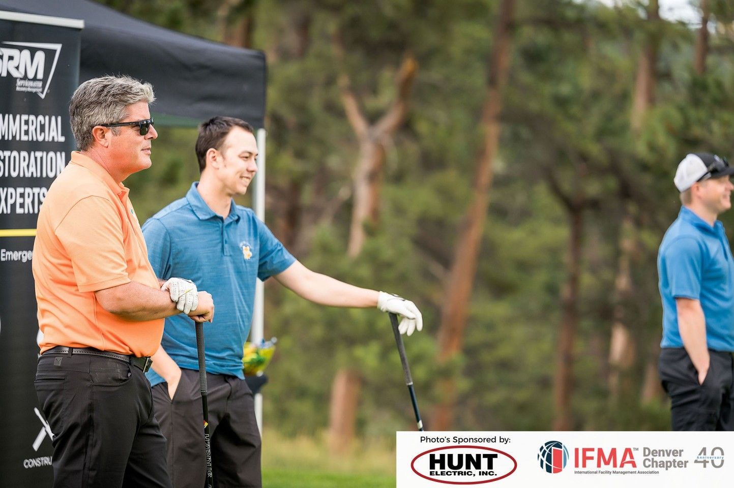 Men in golf attire on a course, looking at something. One points.  A banner is visible.