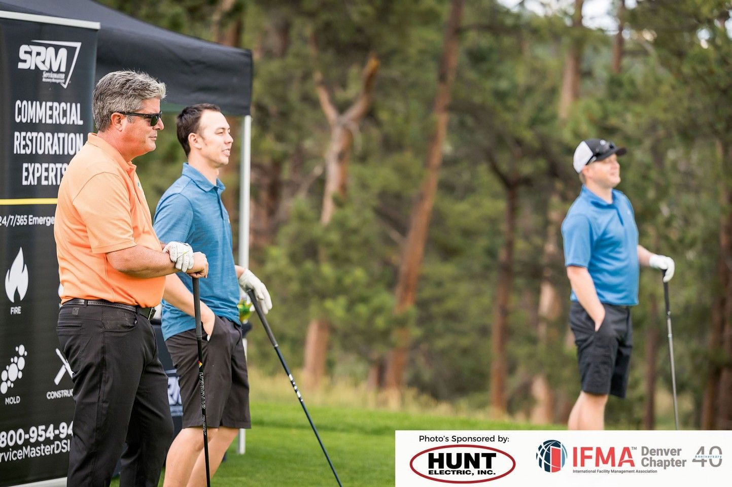 Three men on a golf course: One in orange, two in blue, holding golf clubs, looking at something off-camera.