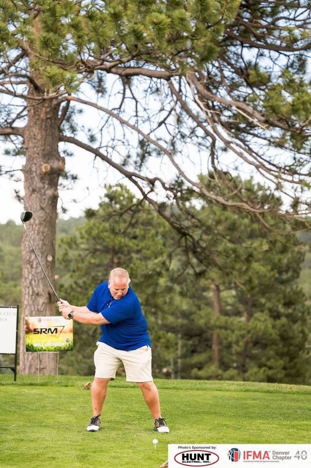 Man swings a golf club on a green, set against a backdrop of trees. He wears blue shirt, khaki shorts, and black shoes.