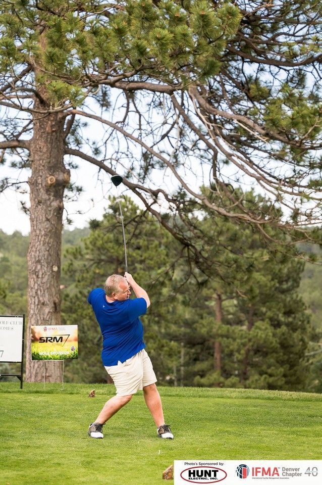 Man in blue shirt swings golf club, tree in the background. Green course, sunny day.