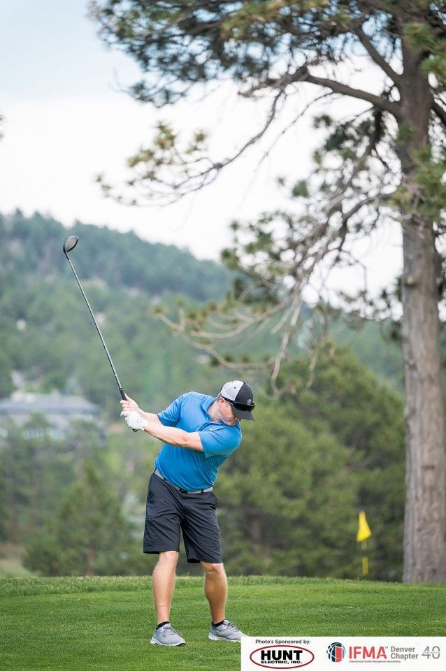Golfer in blue shirt swings club on green course with trees and hills.