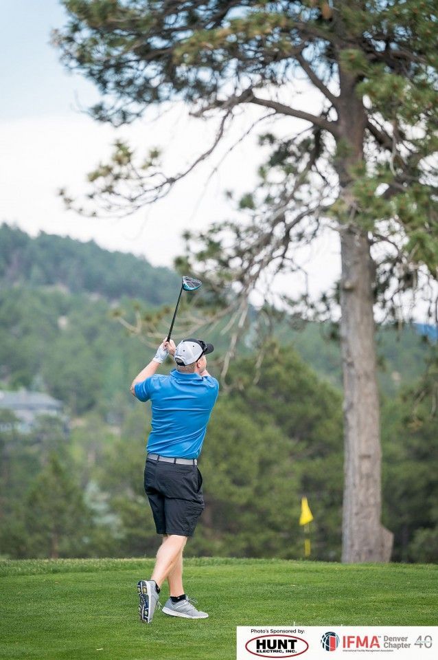 Golfer swinging club on a green course with a tree and mountains in the background.