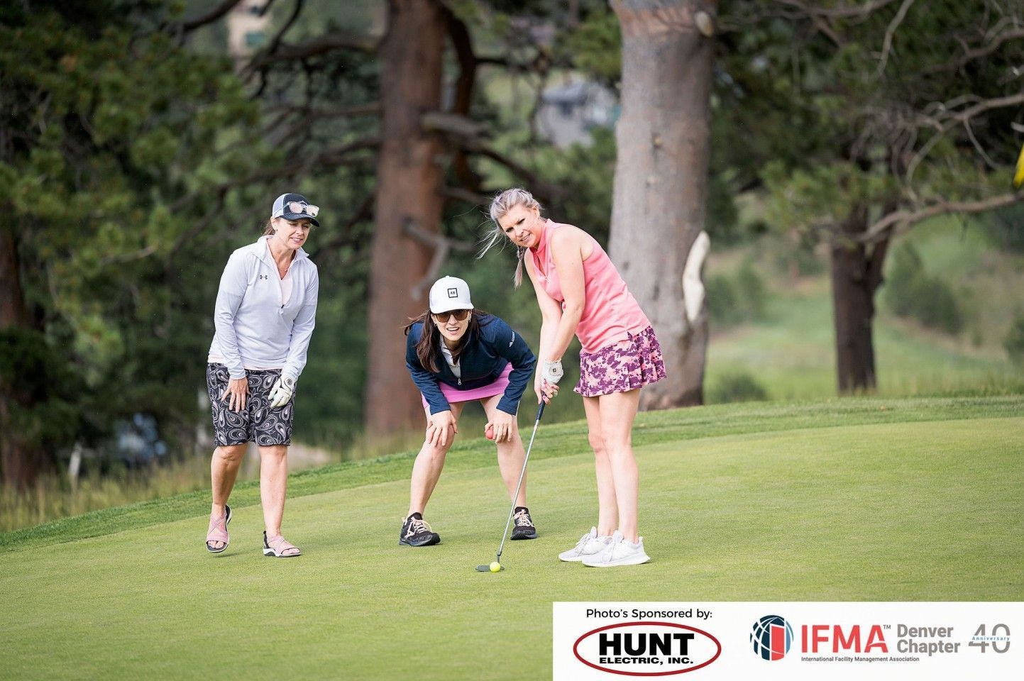 Three women golfing; one prepares to putt, observed by two others on a green, trees in the background.
