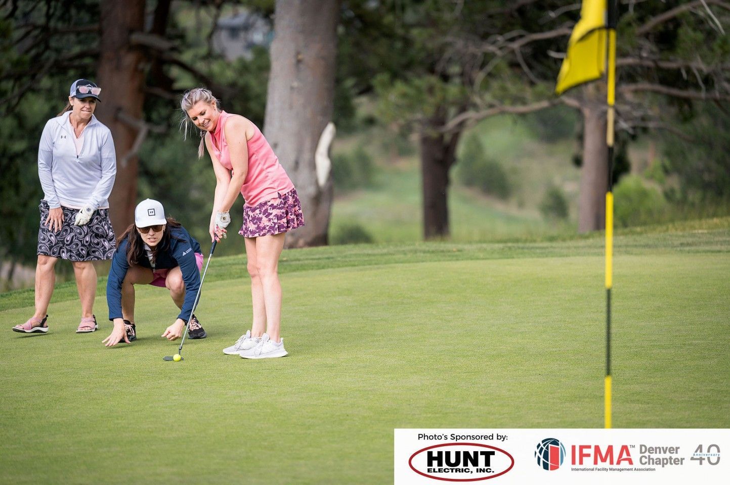 Three women on a golf course: one putting, two watching. Green grass, trees, and a flag.