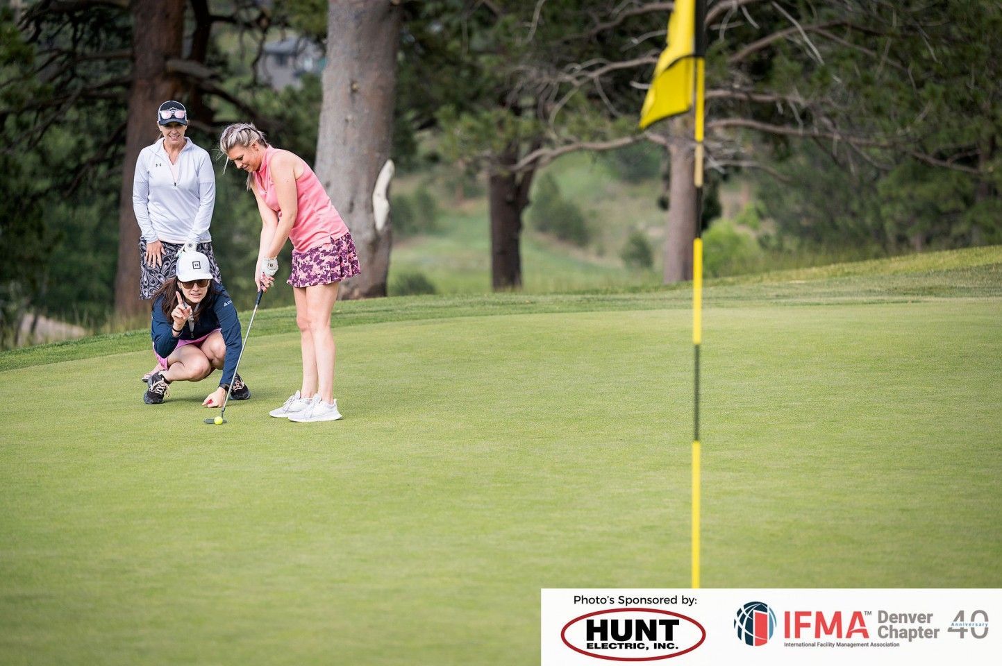 Three women golfing; one puts, two watch on green with flag in the background.