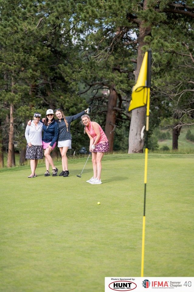 Woman putting golf ball on green while friends pose with flag. Trees and cloudy sky in background.
