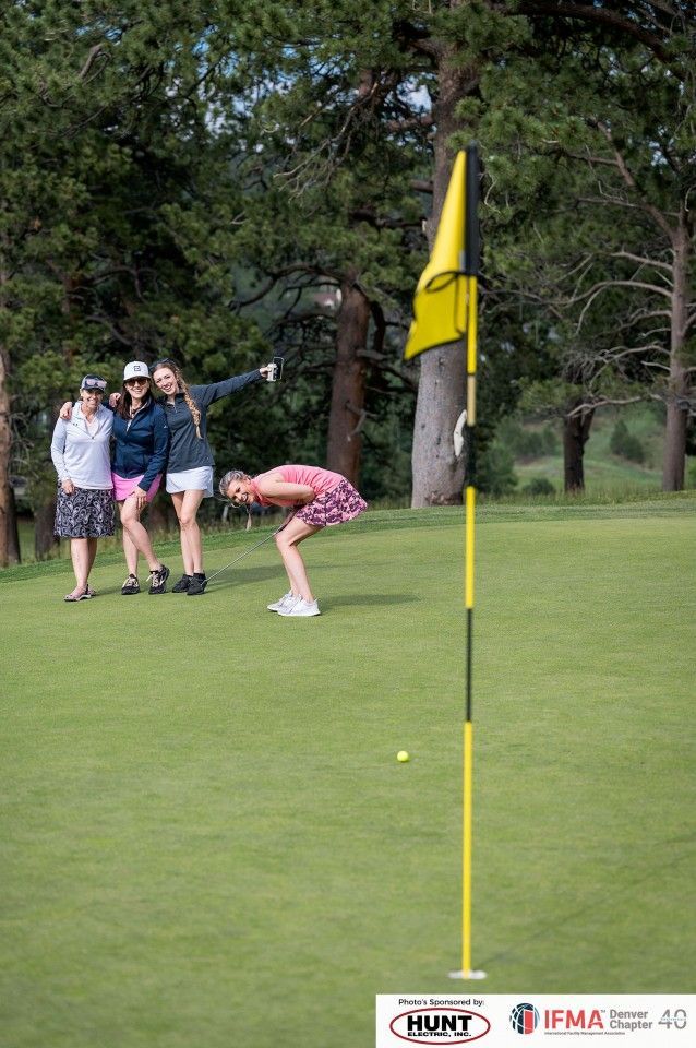 Golfers on green; one bends over ball, others watch and point toward the flag, which is yellow.
