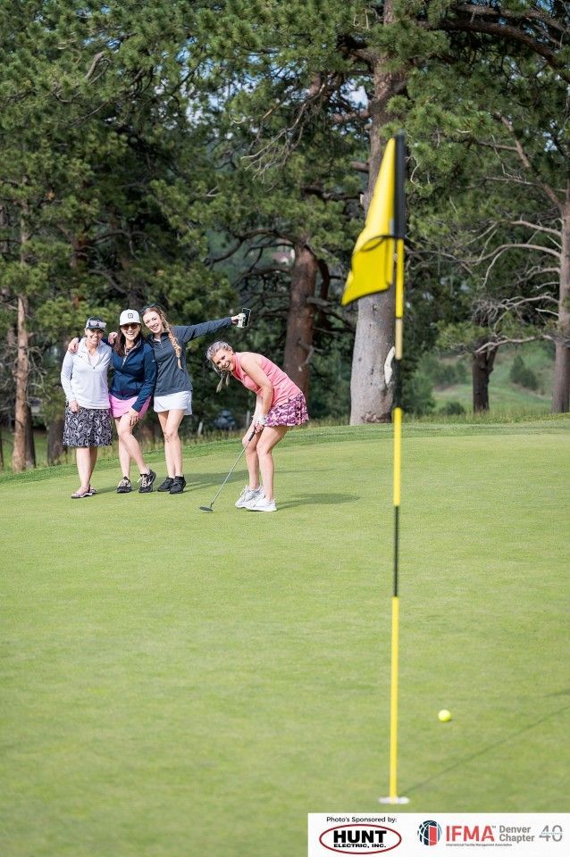 Women golfing, one about to putt, three watching near the green, yellow flag in foreground.