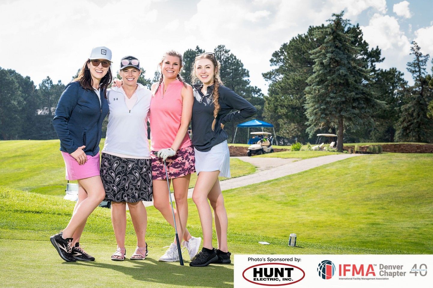 Four women in golf attire pose on a green, smiling. Golf course in background.