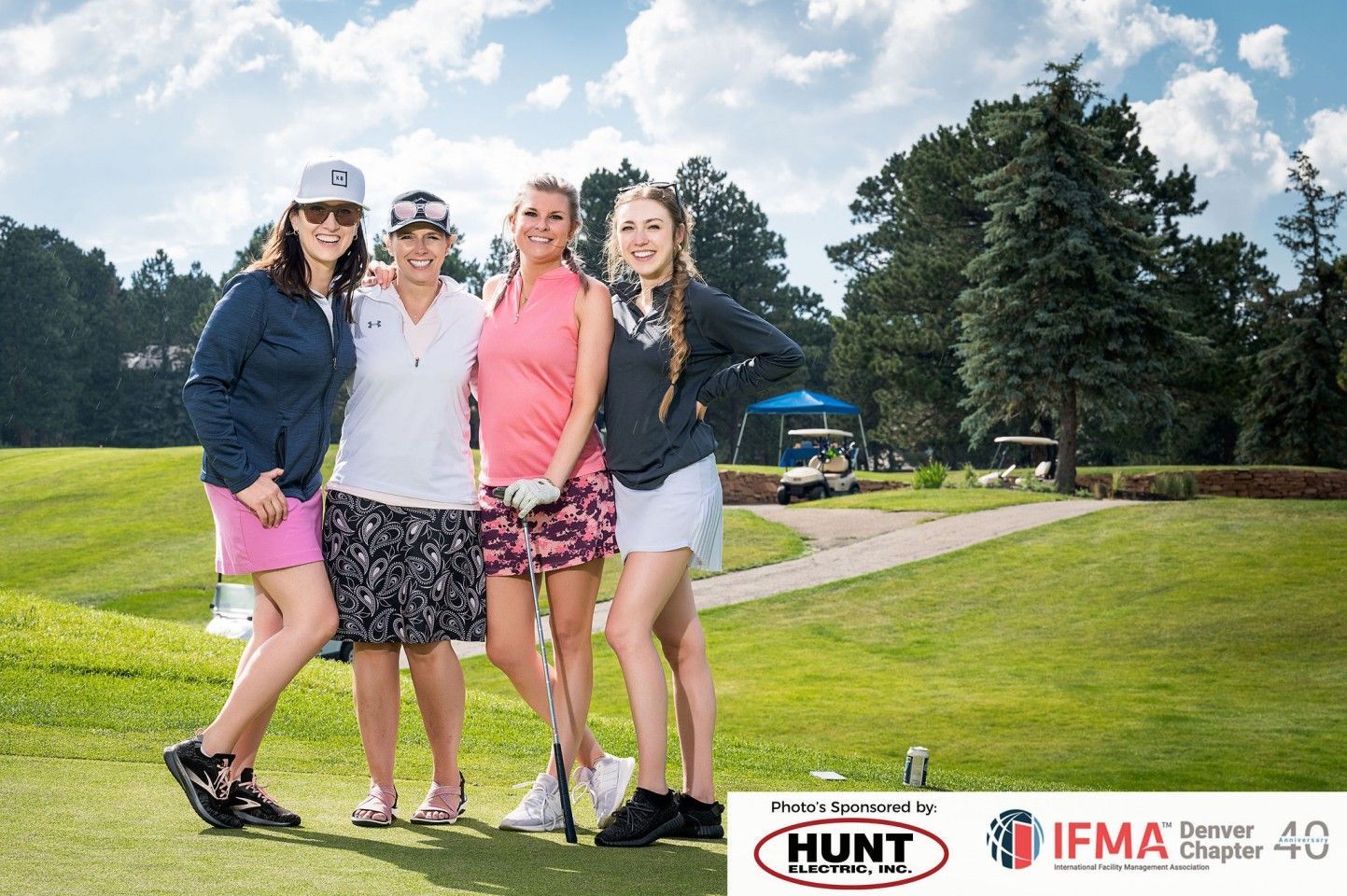 Four women smiling, posing on a golf course with clubs, sunny day.