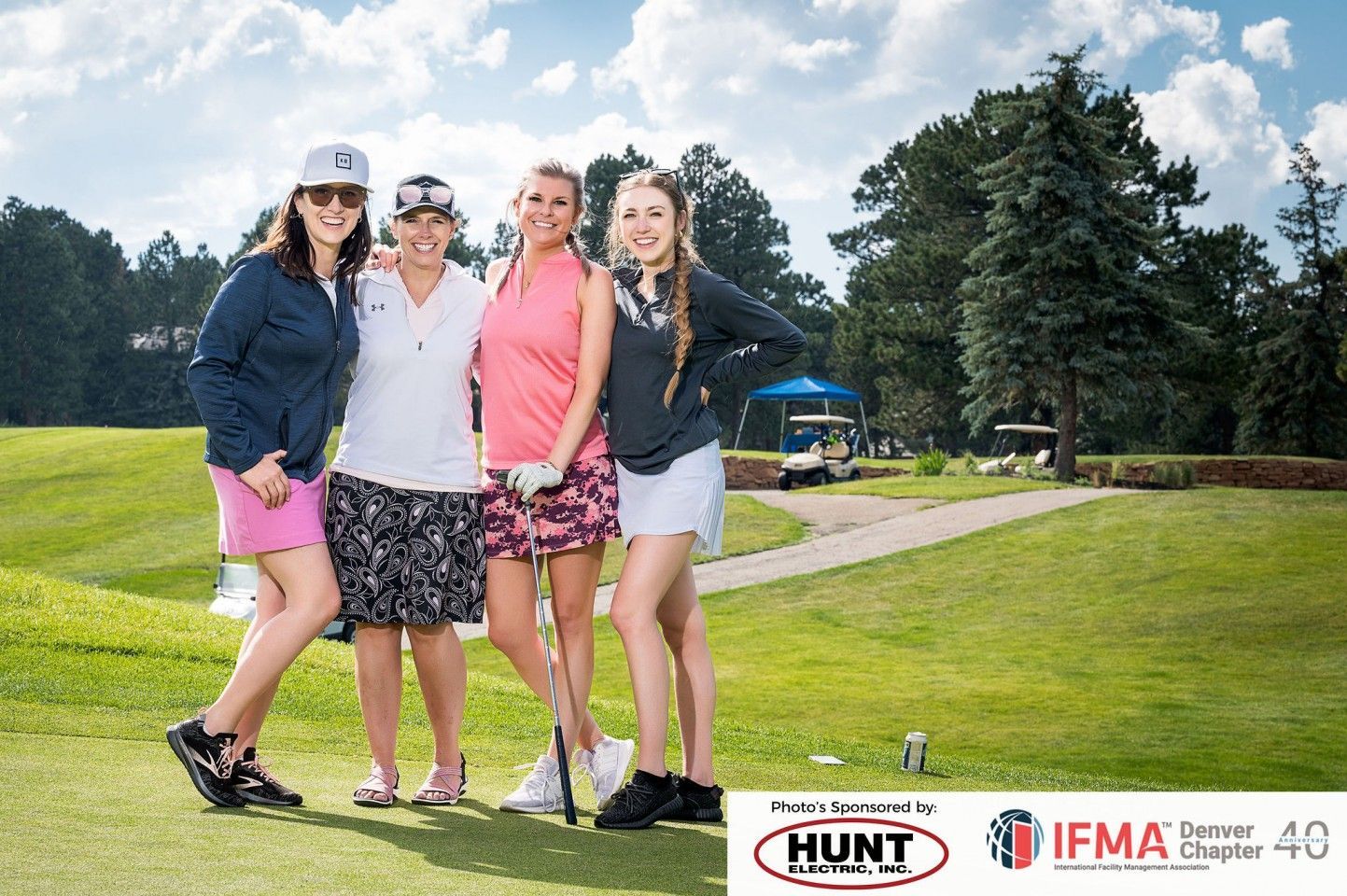 Four women on a golf course posing, smiling. Green grass, blue sky. They wear golf attire.