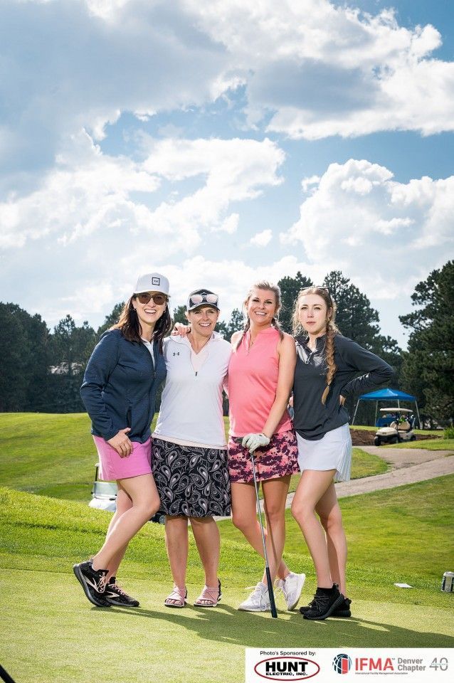 Four women pose on a golf course. They are wearing golf attire, under a blue sky, some holding clubs.